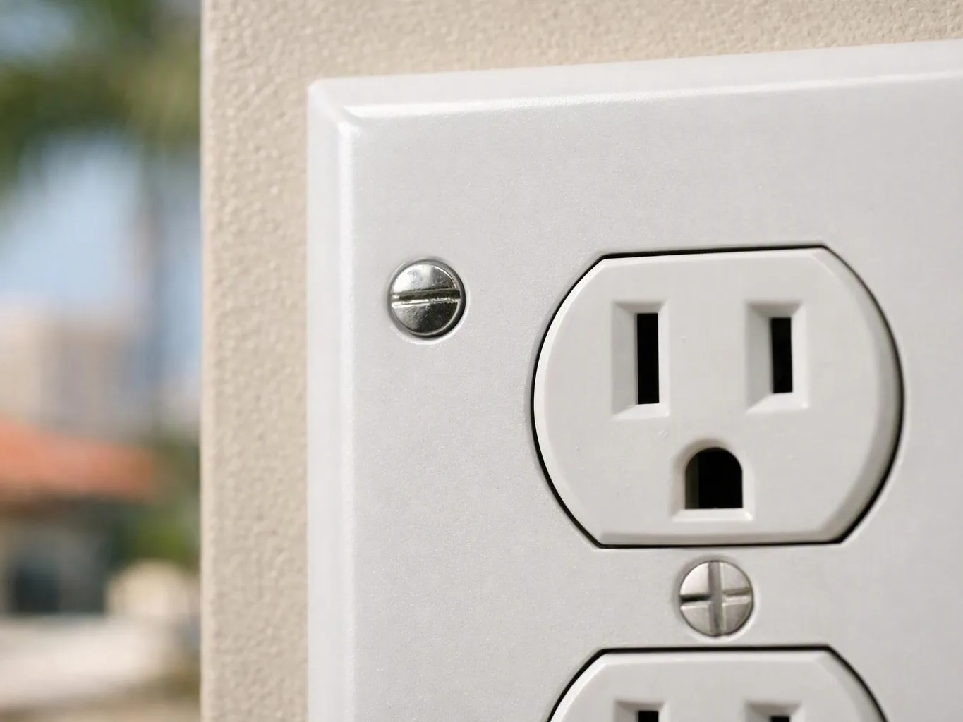 A white electrical wall outlet sits on a beige wall with a blurred outdoor background